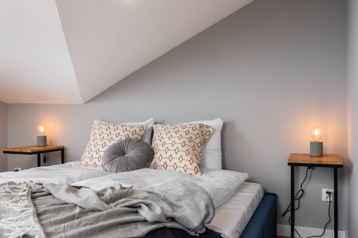 Quarto com teto inclinado, cama com almofadas estampadas, mesas de cabeceira com luminárias e um cobertor na cama.
