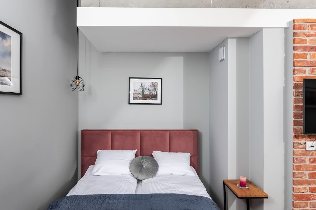 Quarto moderno com cabeceira estofada em vermelho, roupa de cama branca, almofada cinza e uma pequena mesa de cabeceira de madeira com uma vela.