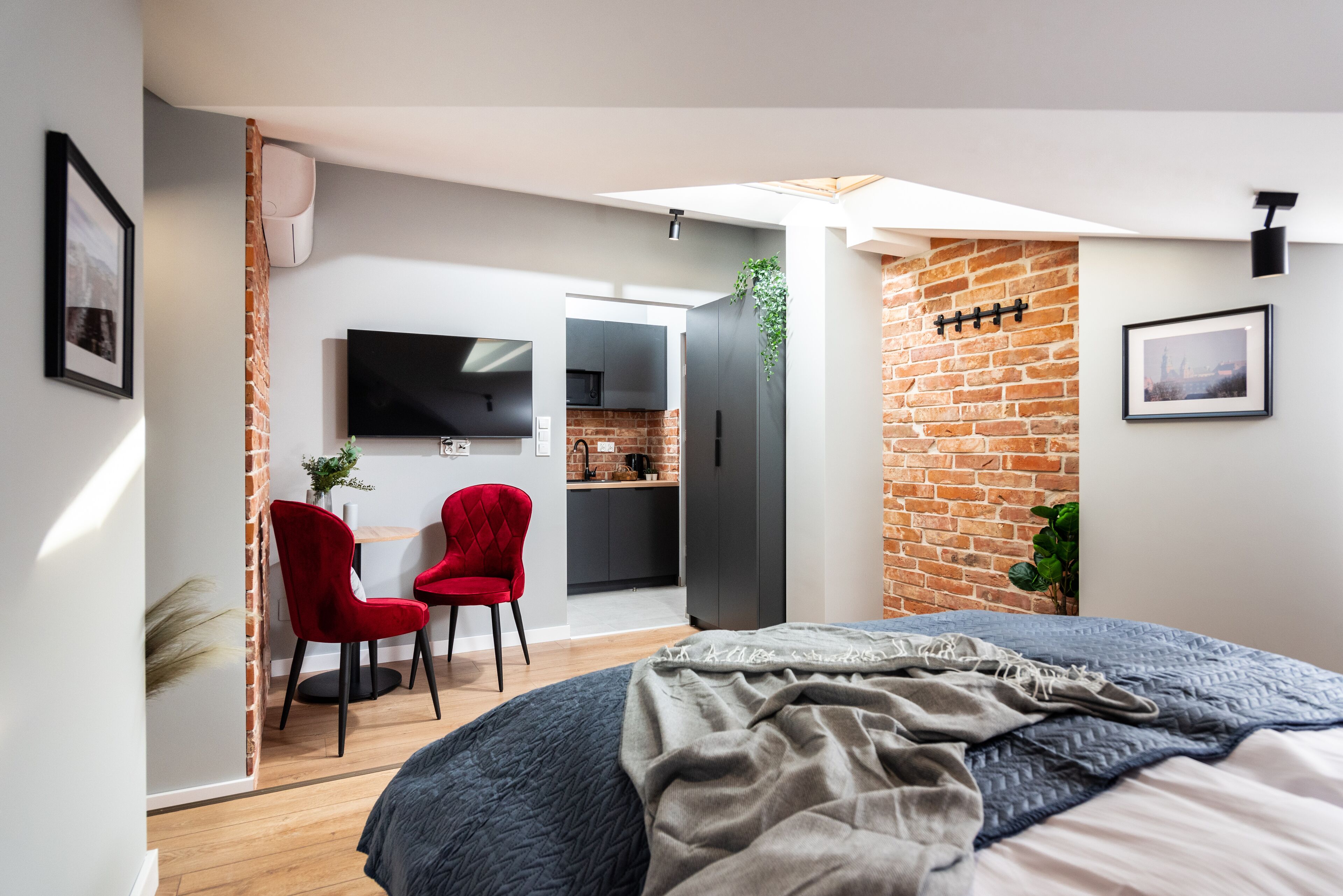 A modern bedroom with a large bed, a red chair, a TV, and a kitchen area visible through an open door.