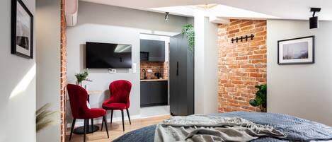 A modern bedroom with a large bed, a red chair, a TV, and a kitchen area visible through an open door.