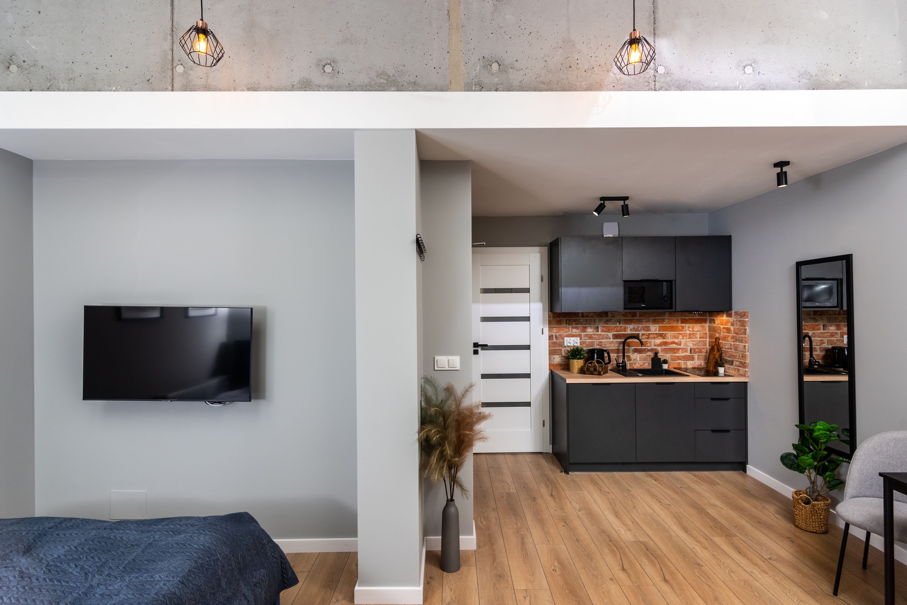 Un espace de vie moderne comprenant un téléviseur à écran plat, un lit, une cuisine avec des meubles sombres et un mur de fond en briques, ainsi qu’un coin repas avec une chaise et une table.