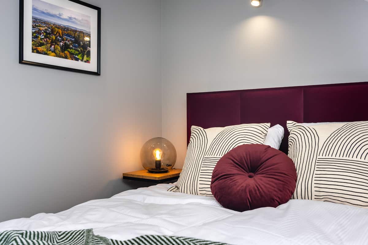 Uma cama bem arrumada com roupa de cama listrada, um travesseiro vermelho e uma mesa de cabeceira com abajur.