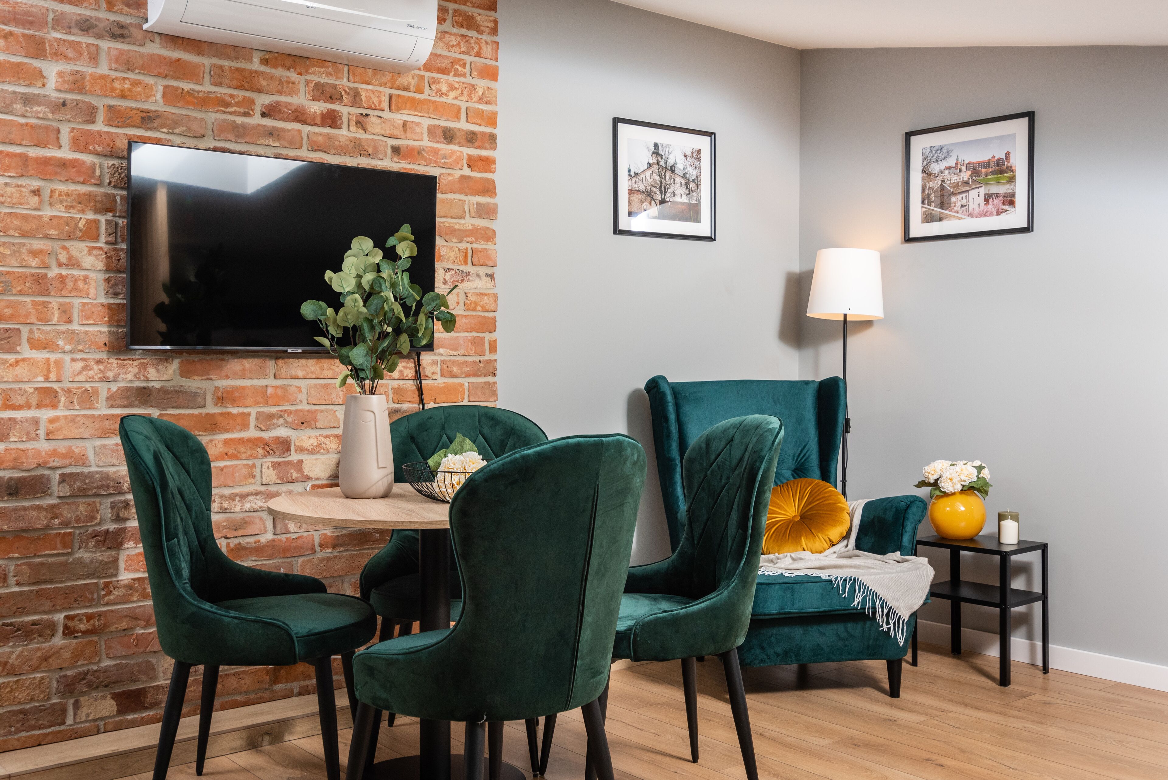 Una sala de estar moderna con una pared de ladrillo, un televisor de pantalla plana, sillas color turquesa, una mesa redonda de madera y cuadros en la pared.