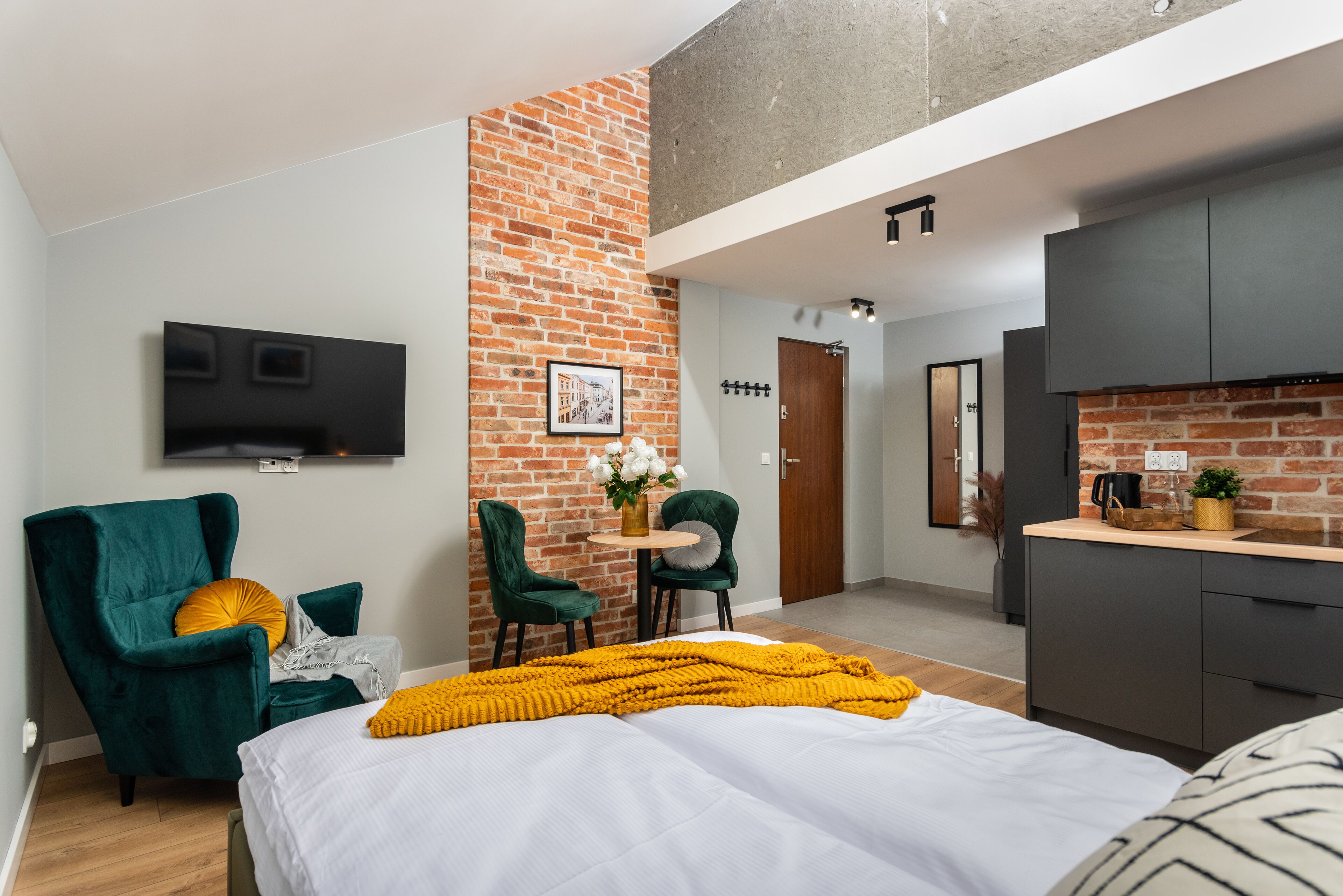 Ett modernt hotellrum med en accentvägg i tegel, en platt-TV som hänger ovanför en grön stol, ett litet runt bord med stolar, en säng med ett vitt och gult överkast och ett litet pentry med mörka skåp.
