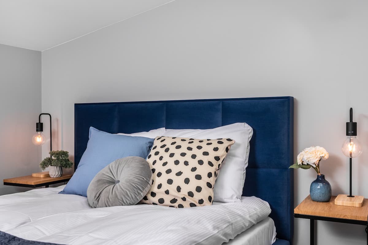Uma cama bem arrumada com cabeceira azul, uma mesinha de cabeceira com uma planta e um abajur.