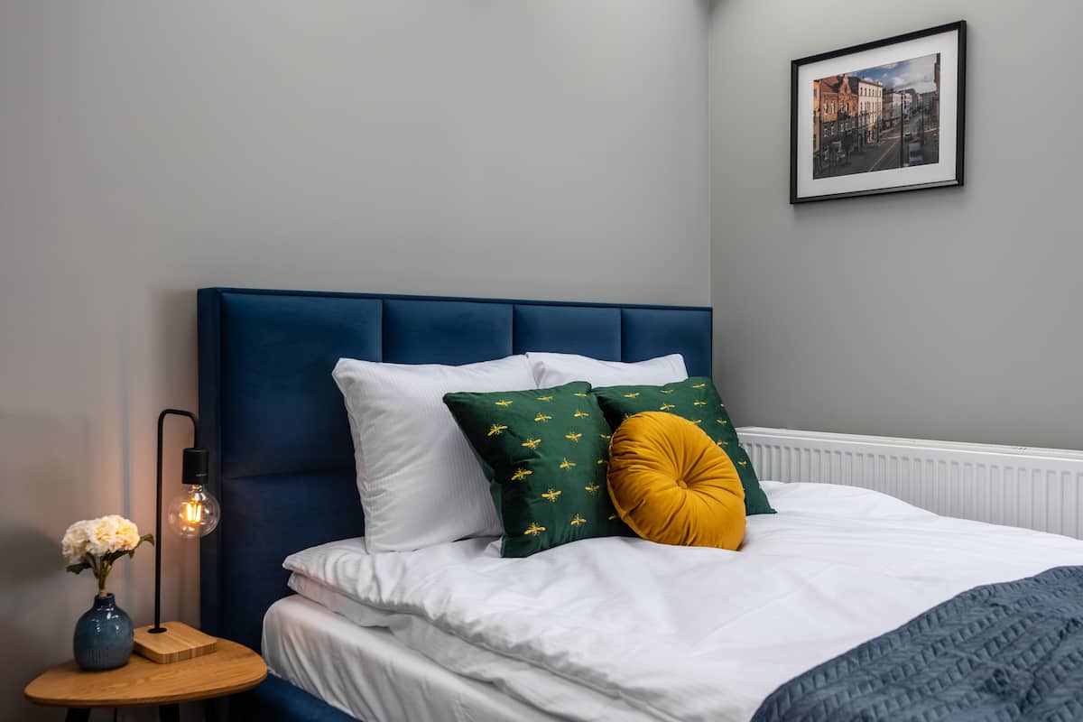 Cama bem arrumada com lençóis brancos, cabeceira azul e almofadas decorativas em tons de verde e mostarda.