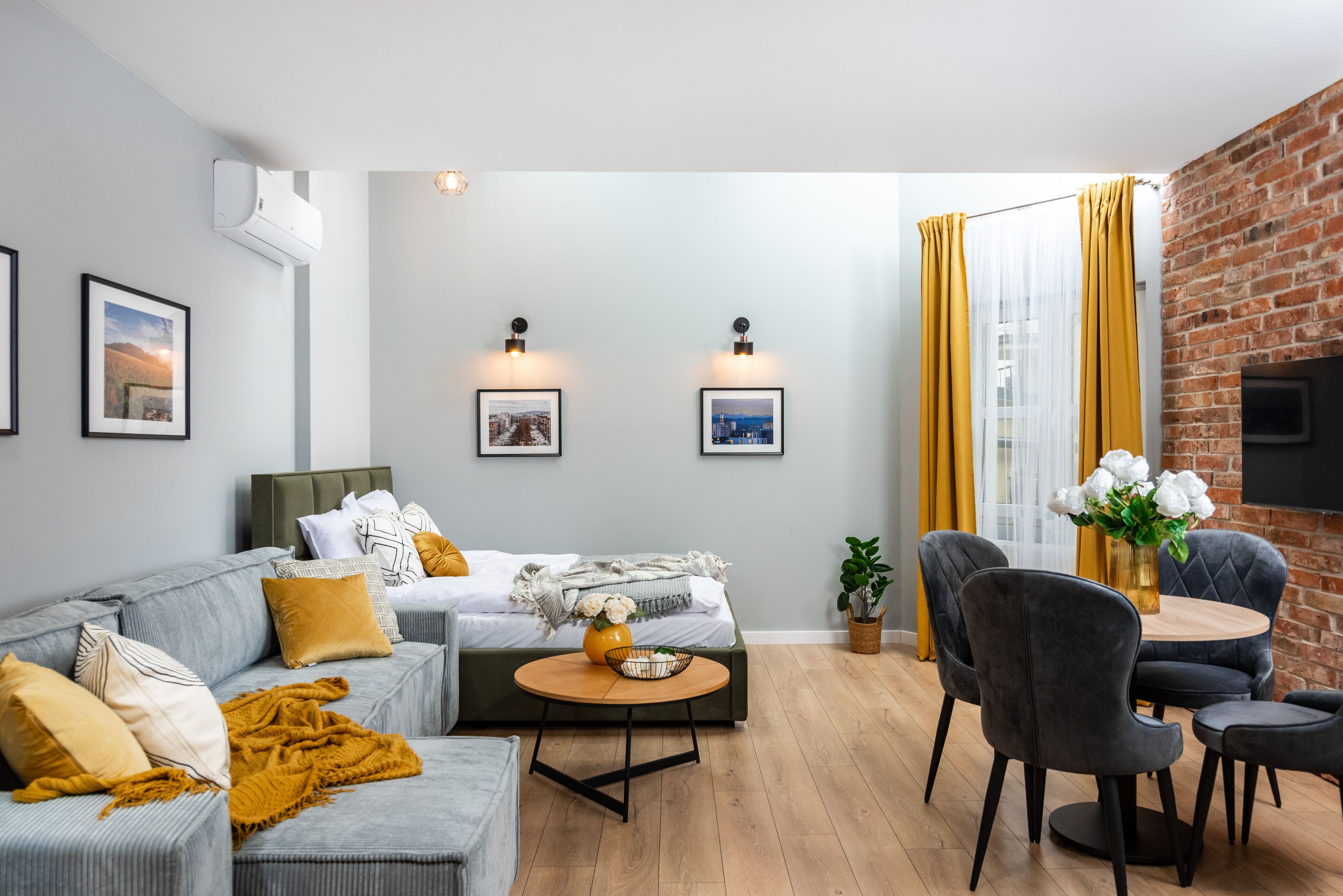 Una sala de estar moderna con un sofá gris, una mesa de centro redonda y una zona de comedor con pared de ladrillo y cortinas amarillas.
