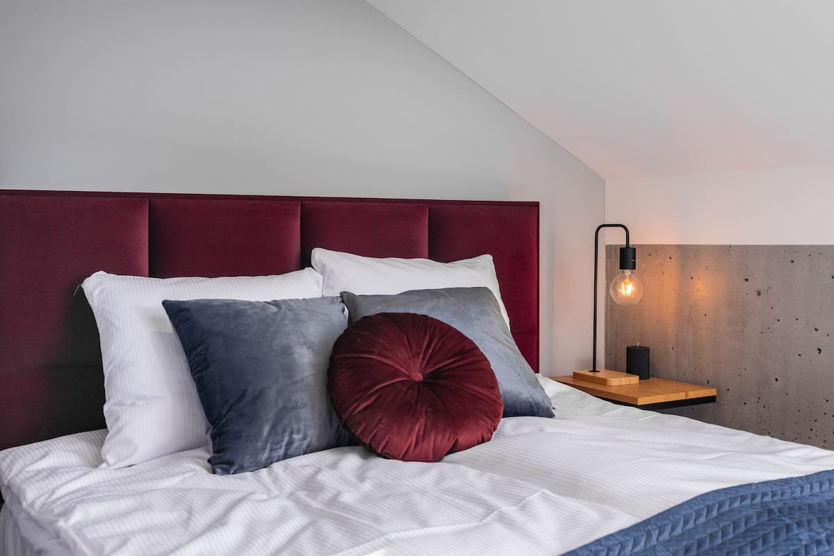 Cama bem arrumada com cabeceira vermelha, travesseiros brancos e cinzentos e um cobertor azul. Há uma mesinha de cabeceira com abajur e uma pequena prateleira de madeira.