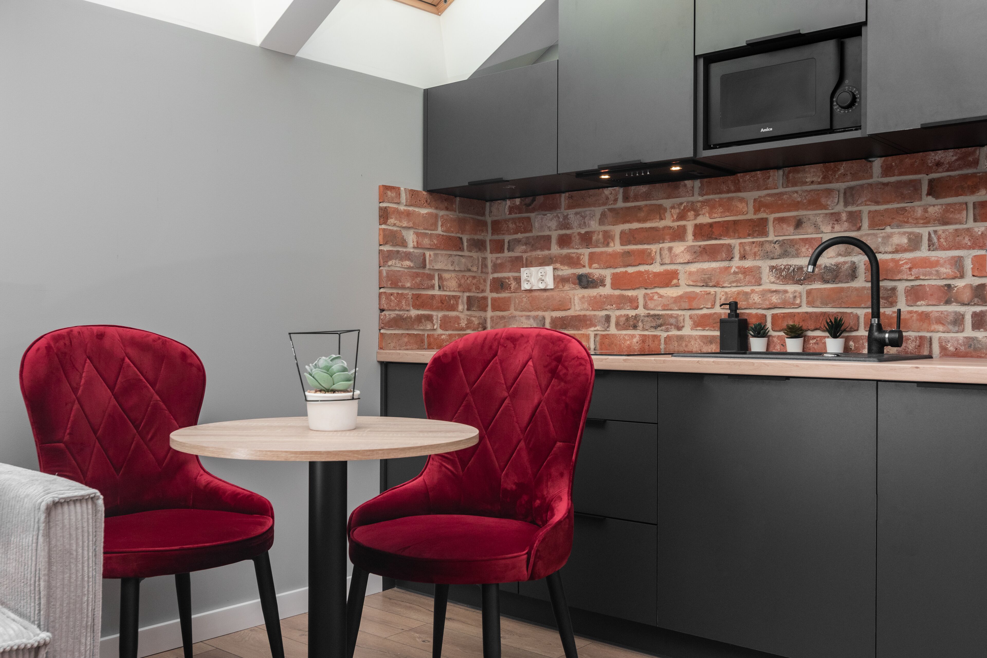 A compact kitchen with a brick backsplash, two red velvet chairs, a small round table, and a modern microwave.