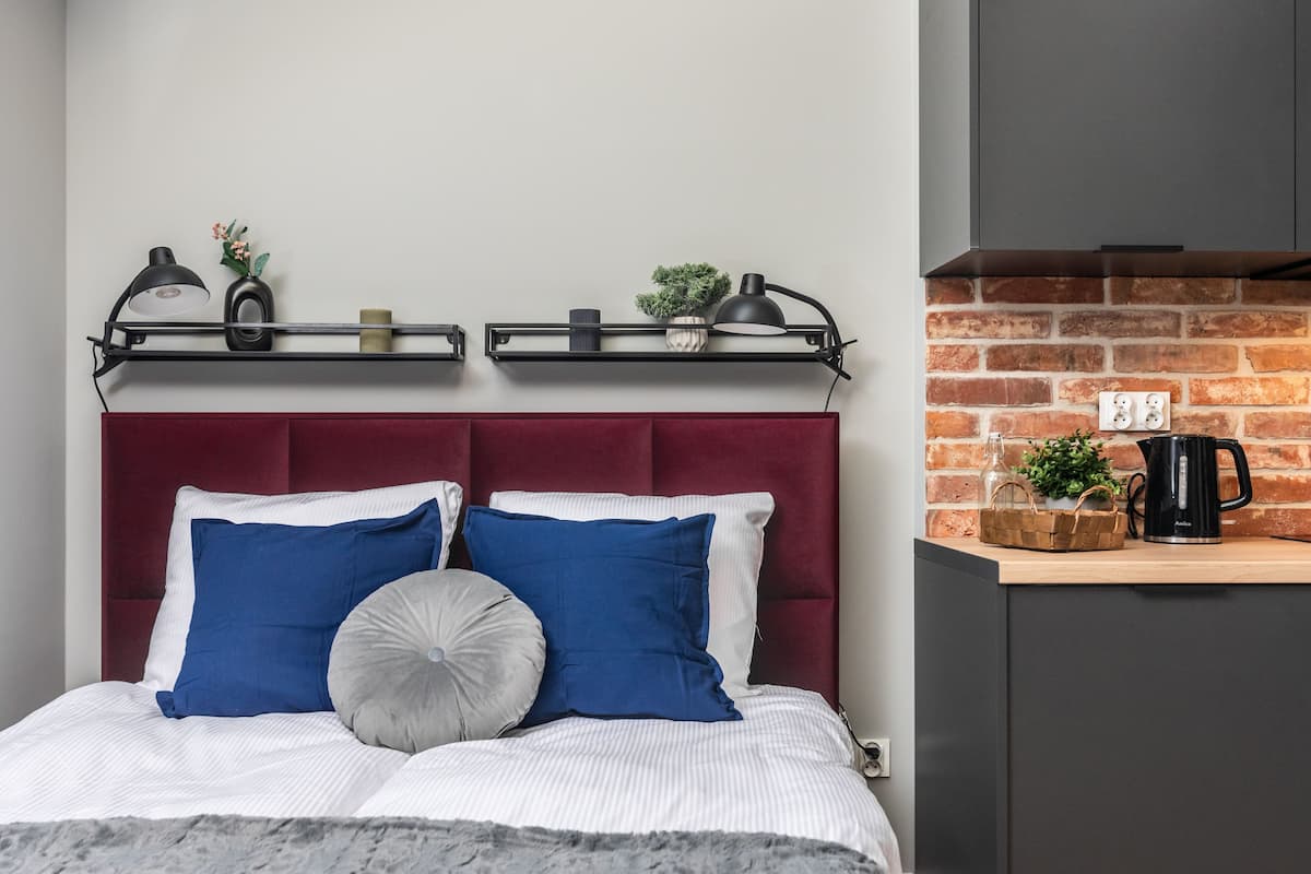Uma cama com cabeceira vermelha, almofadas azuis e brancas e uma almofada cinzenta. Acima da cama, duas prateleiras exibem objetos decorativos e um abajur. À direita, há uma área de cozinha com parede de tijolos, uma cafeteira e um cesto com plantas.