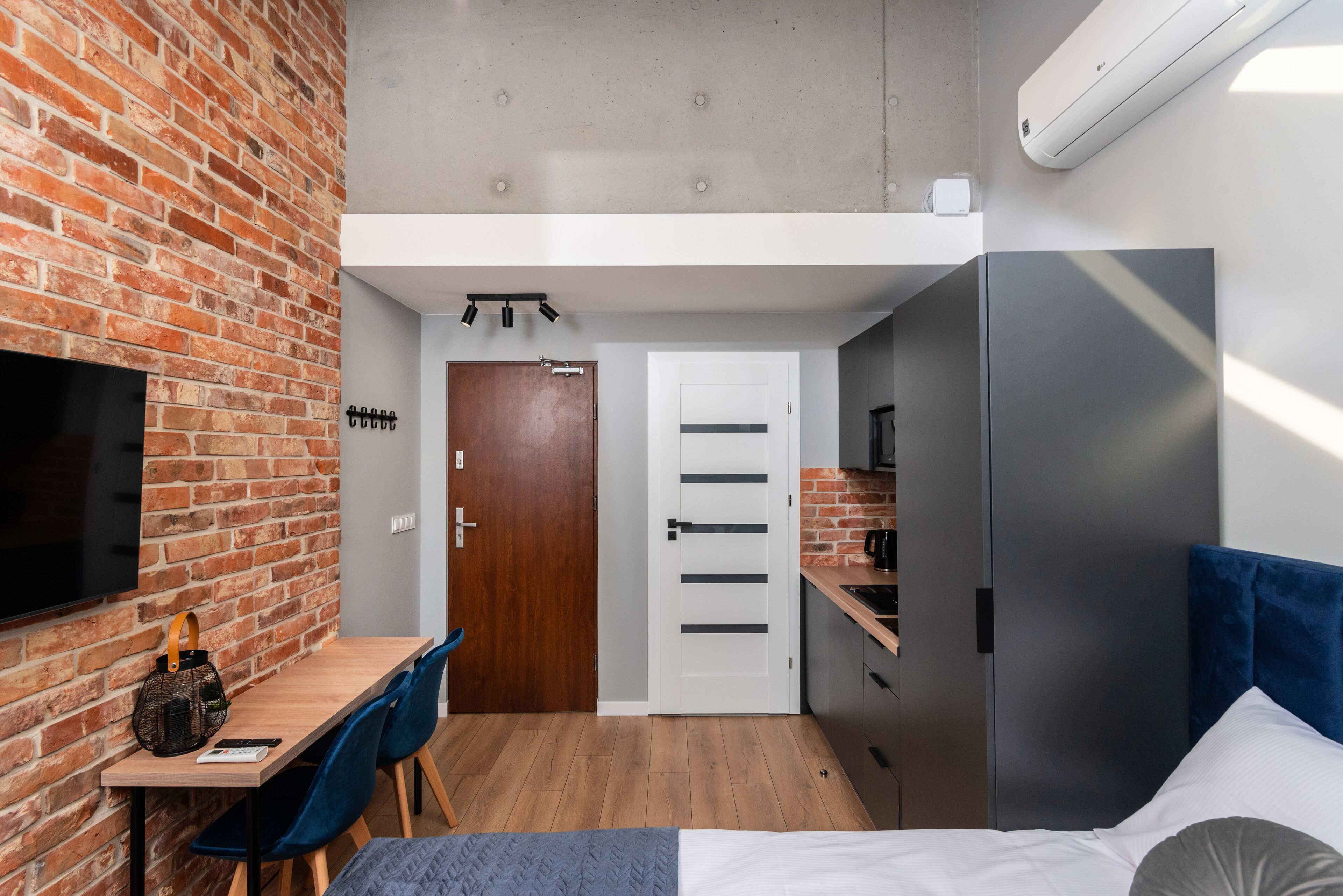 Una habitación pequeña con pared de ladrillo, un escritorio de madera, una cama con cabecera azul y una kitchenette.