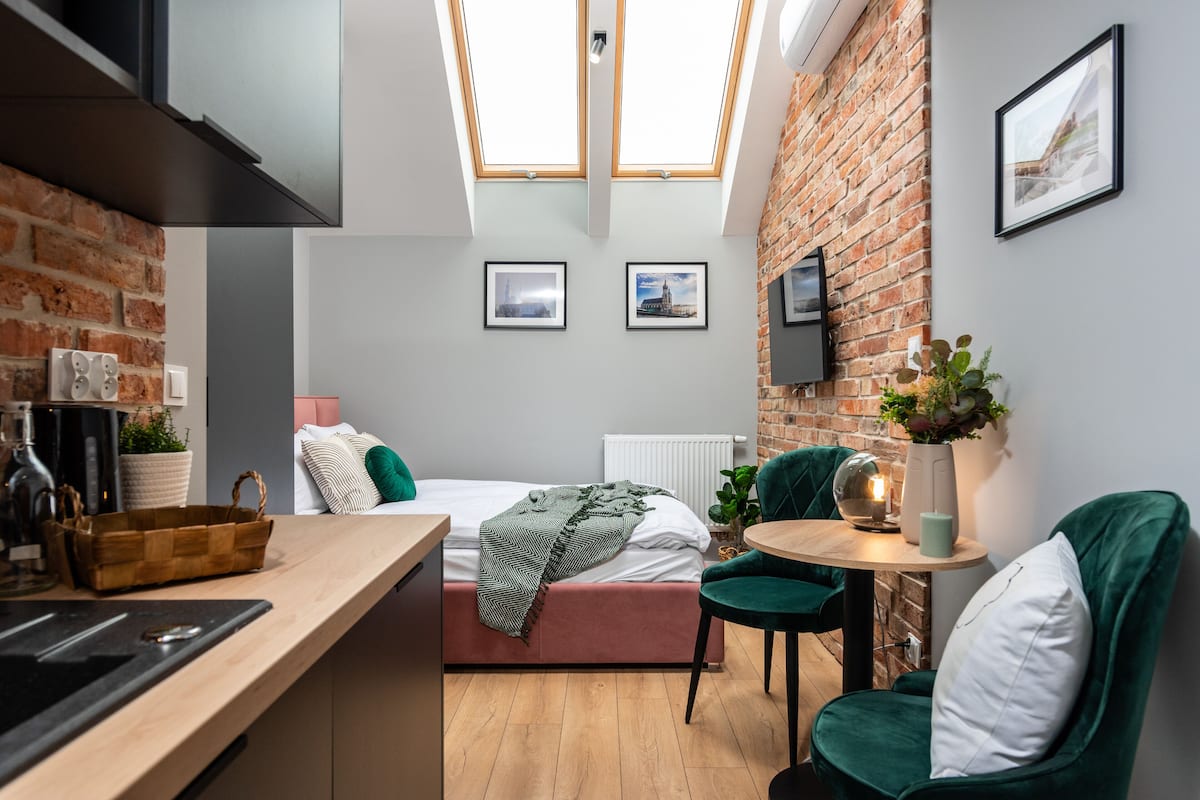 Quarto compacto com cozinha compacta, uma cama com manta verde, pequena área de jantar com mesa e cadeiras, e parede de tijolos com duas claraboias.