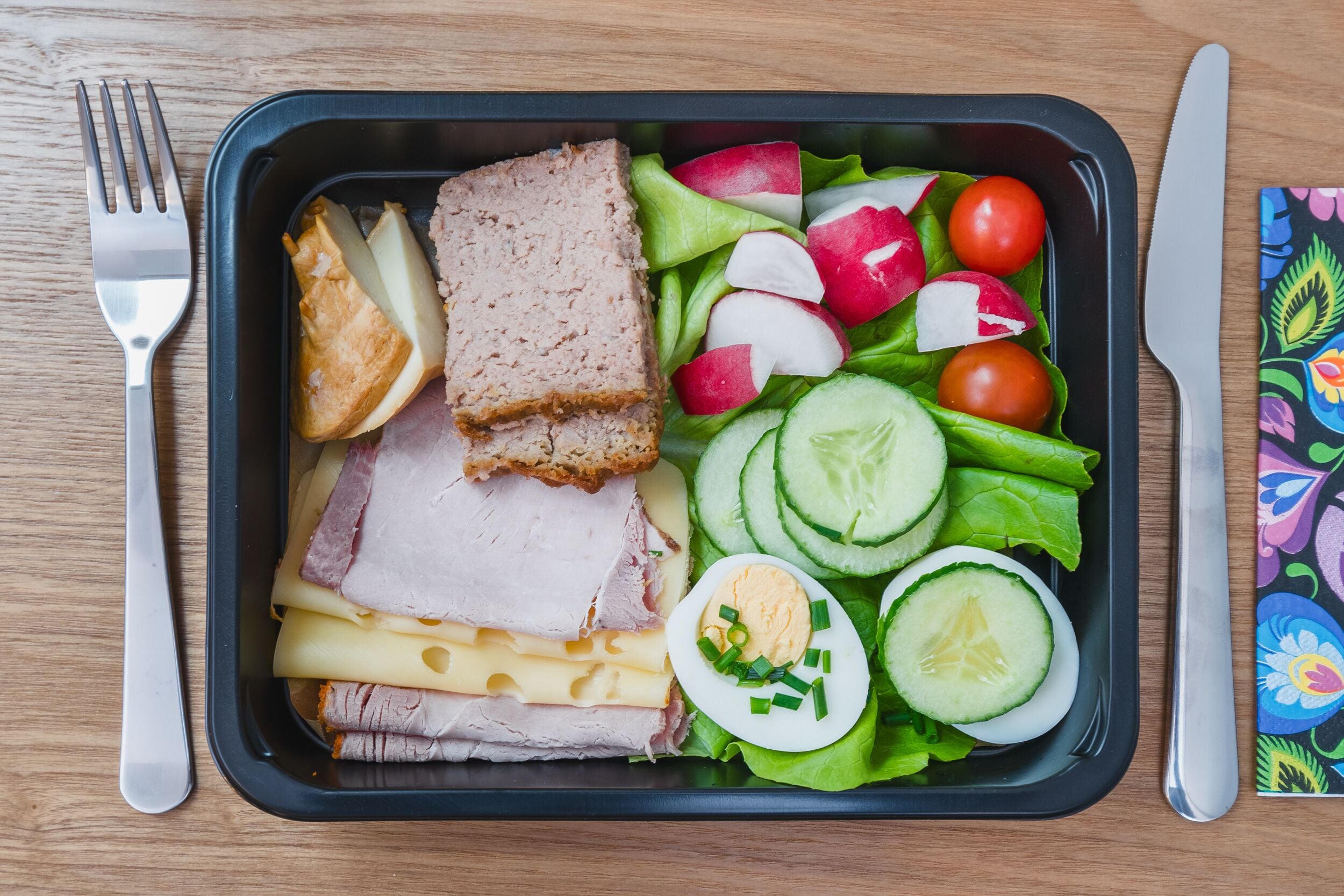 A meal in a black tray with ham, cheese, eggs, and a salad of cucumbers, radishes, and tomatoes.