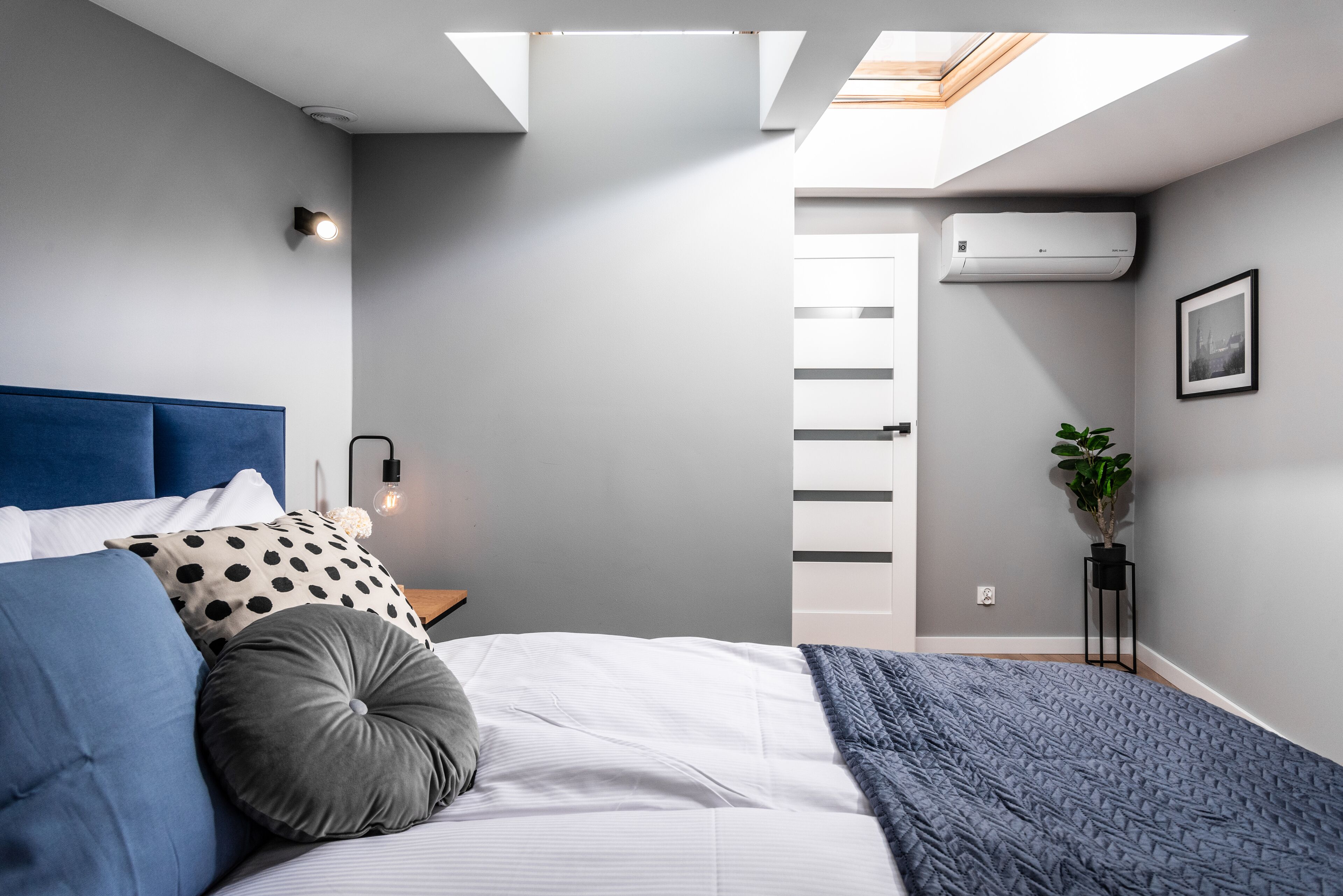 Ein modernes Schlafzimmer mit Dachfenster, einem Bett mit blau-grauen Betttextilien, einem Nachttisch mit Lampe, einer wandmontierten Klimaanlage und einer Zimmerpflanze.