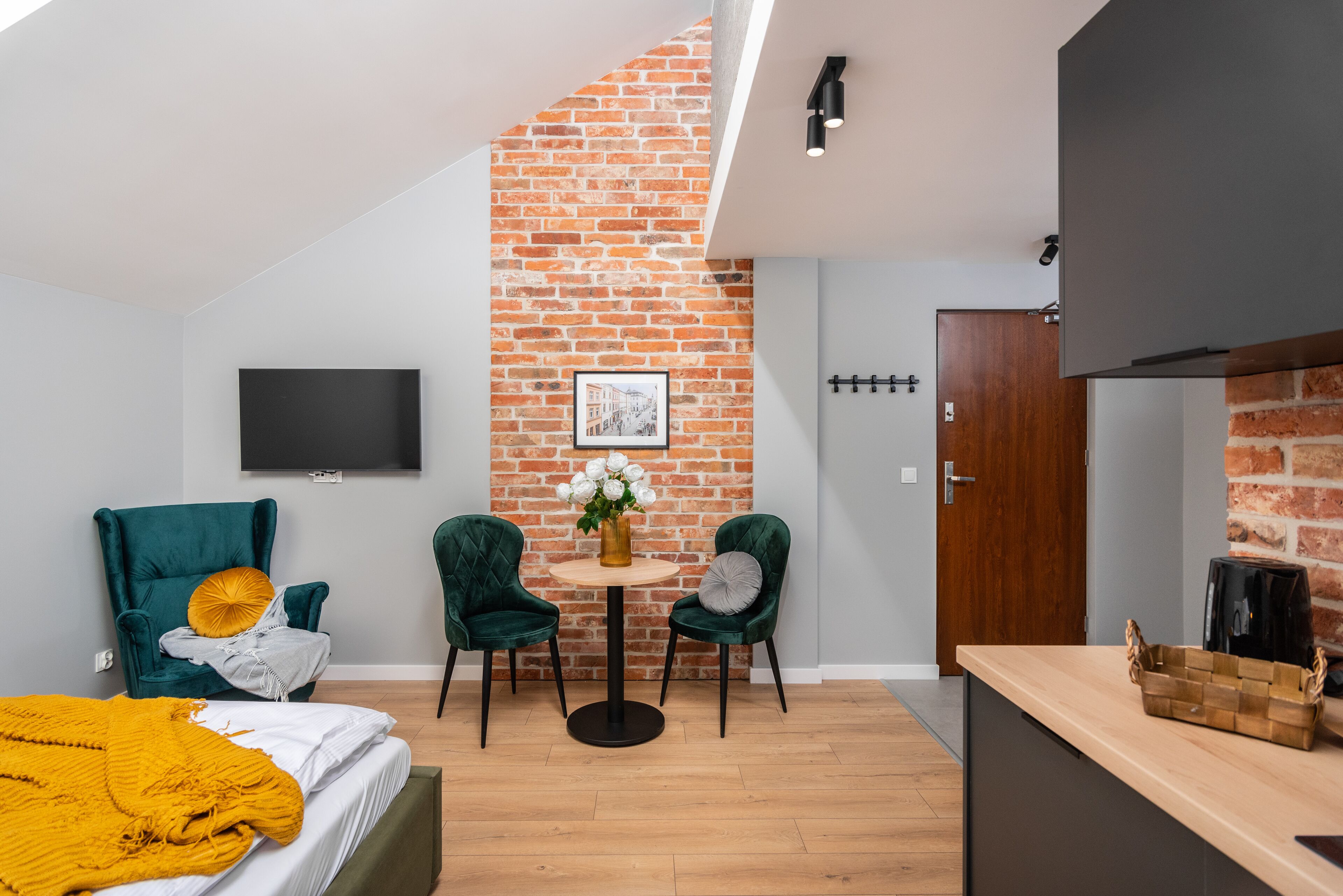 A modern room with a brick wall, a flat-screen TV, two green chairs, a small round table, and a bed with a yellow blanket.