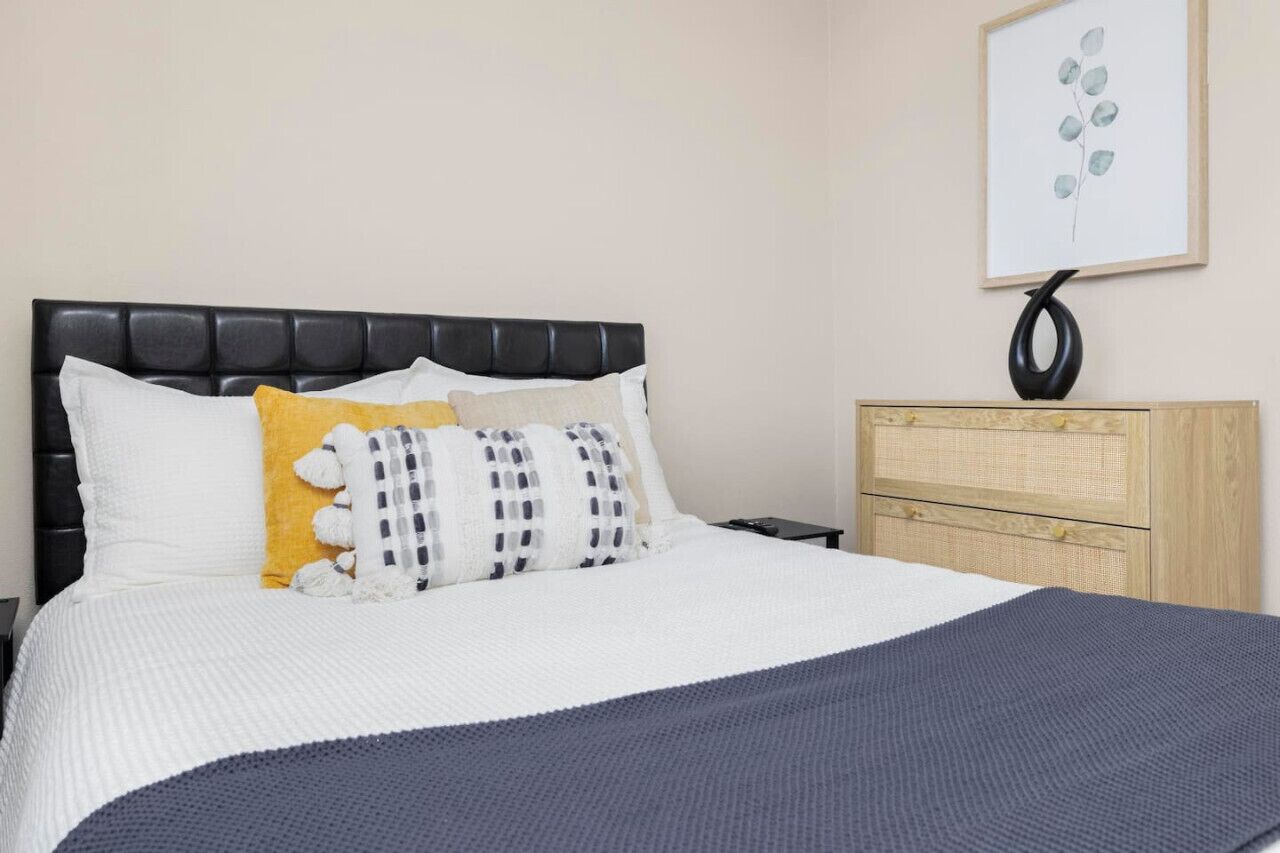 A neatly made bed with white and gray bedding, a yellow pillow, and a white stuffed toy. There is a wooden nightstand with a black vase and a framed picture of leaves on the wall.