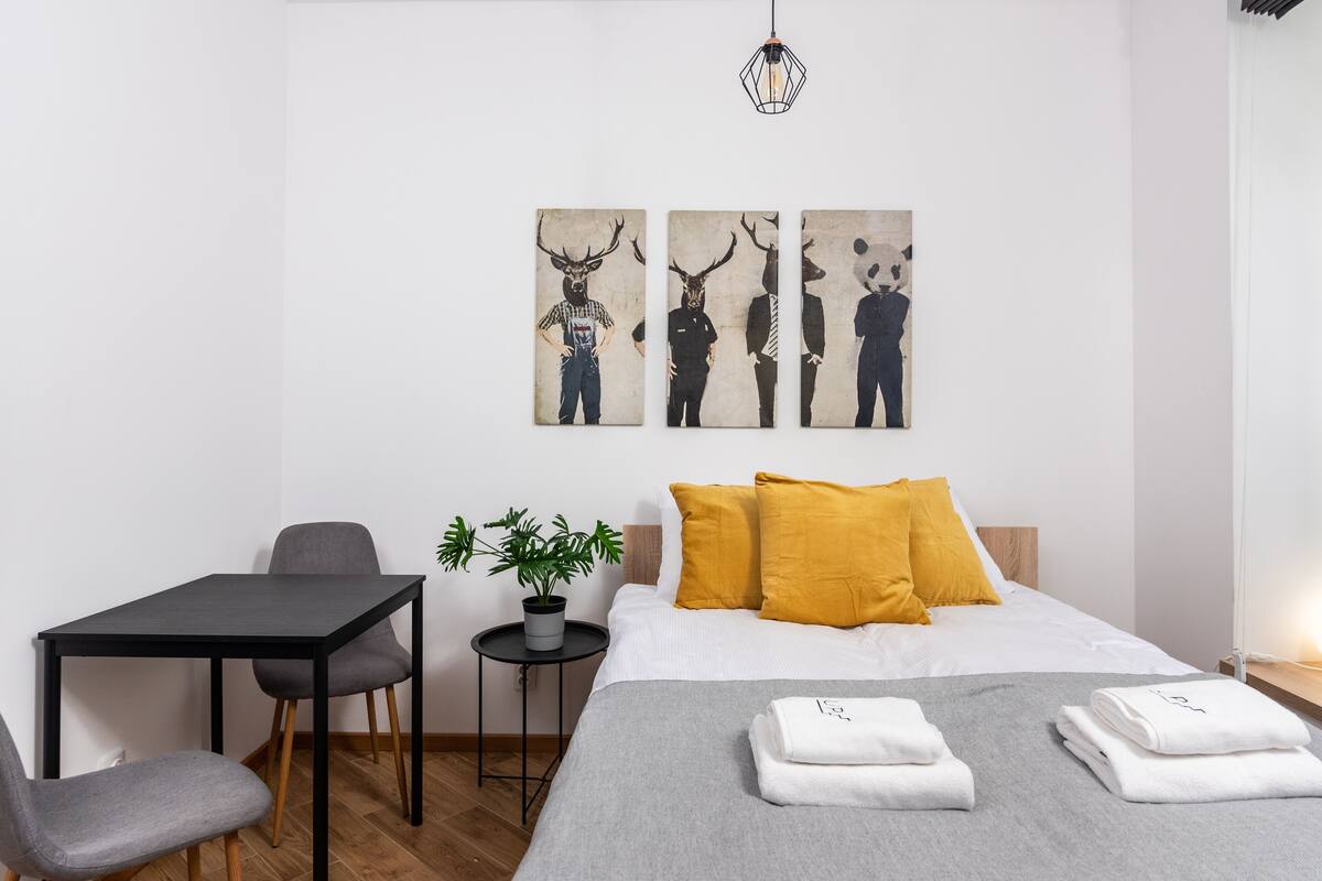 A modern hotel room with a bed, a small table with two chairs, a potted plant, and artwork on the wall.
