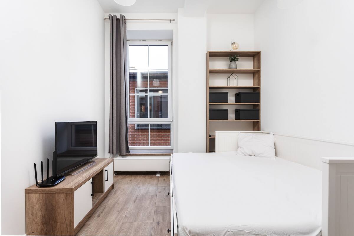 A modern bedroom with a bed, a TV on a wooden console, a window with a view of a brick building, and a built-in shelving unit.