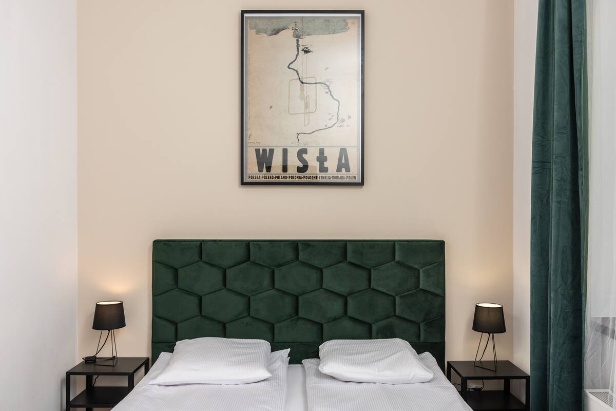A hotel room with a green upholstered headboard, two white pillows, and two bedside tables with lamps.