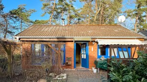 Un bâtiment d’un seul étage, avec un toit recouvert de tuiles, une porte bleue et une petite construction attenante surmontée d’une auvent blanche.