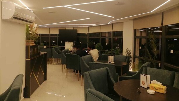 A modern restaurant with a central dining area, surrounded by high-backed chairs and tables, with a flat-screen TV mounted on the wall.