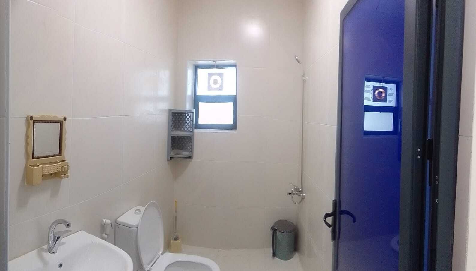 Ein kleines Badezimmer mit Toilette, Waschbecken und Dusche. Ein Fenster bietet Blick ins Freie, eine Mülleimer steht bereit und eine Tür mit Griff ist vorhanden.