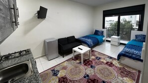 A compact living space with a kitchenette, two beds with blue bedding, a small white table, and a colorful rug.