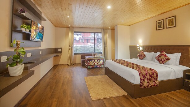 Habitación de hotel con techo de madera, cama grande con ropa blanca, mesita de noche con lámpara, escritorio pequeño con silla, ventana con cortinas y estante en la pared con plantas.