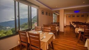 Comedor con mesas dispuestas para dos, ventanas amplias con vistas a montañas y cuadros en las paredes.