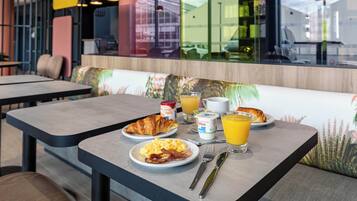 A breakfast setup with croissants, scrambled eggs, bacon, orange juice, and yogurt on a table in a modern dining area with a view through a glass window.