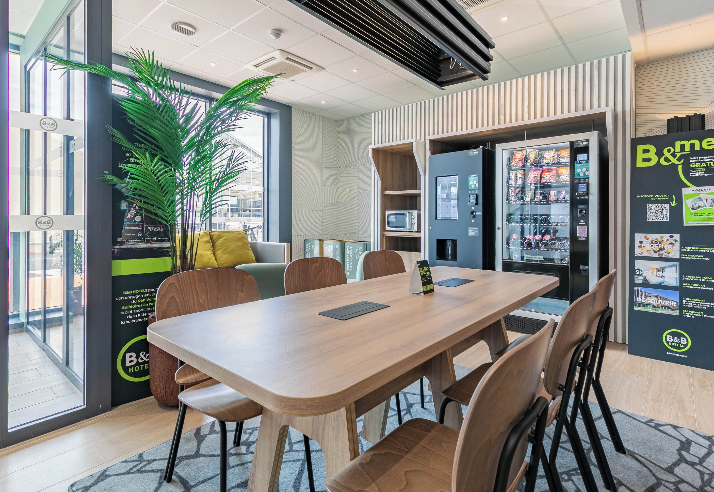 A modern hotel room with a wooden dining table, comfortable seating, a large potted plant, a vending machine, and a view of the city through large windows.