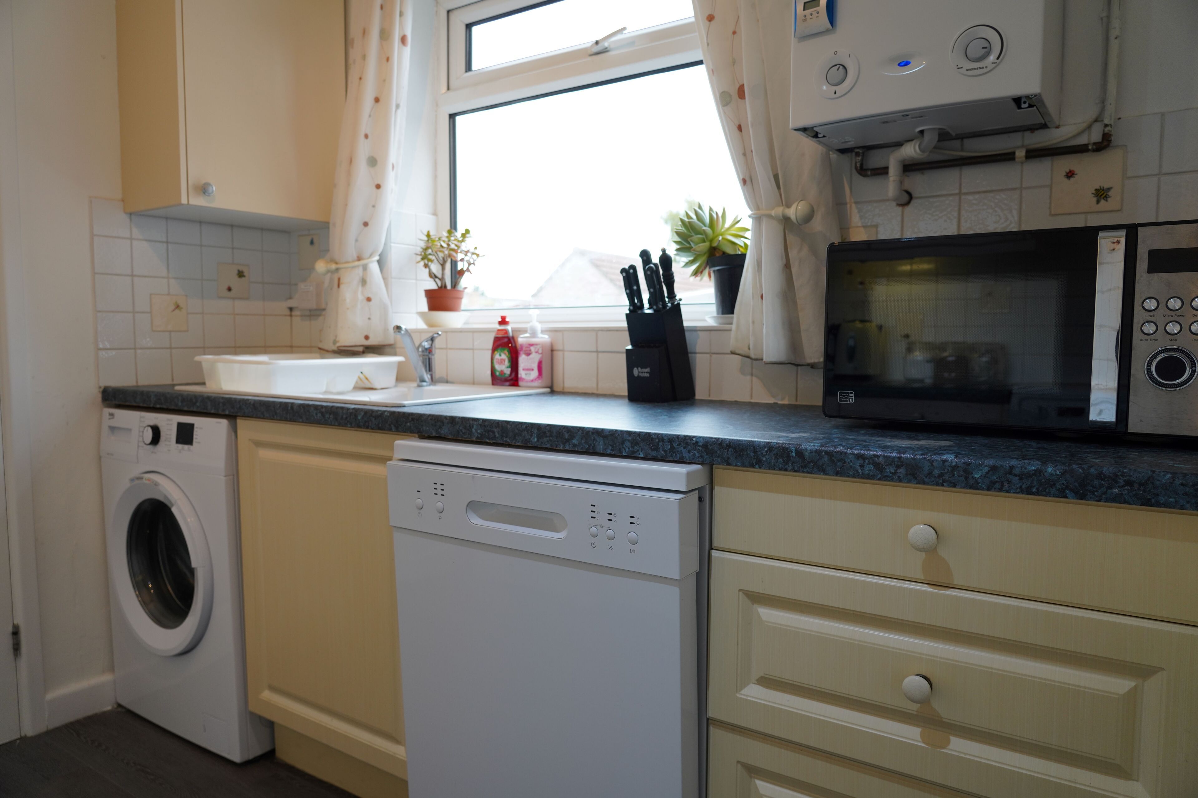 A kitchen with a washing machine, dishwasher, microwave, and potted plants.