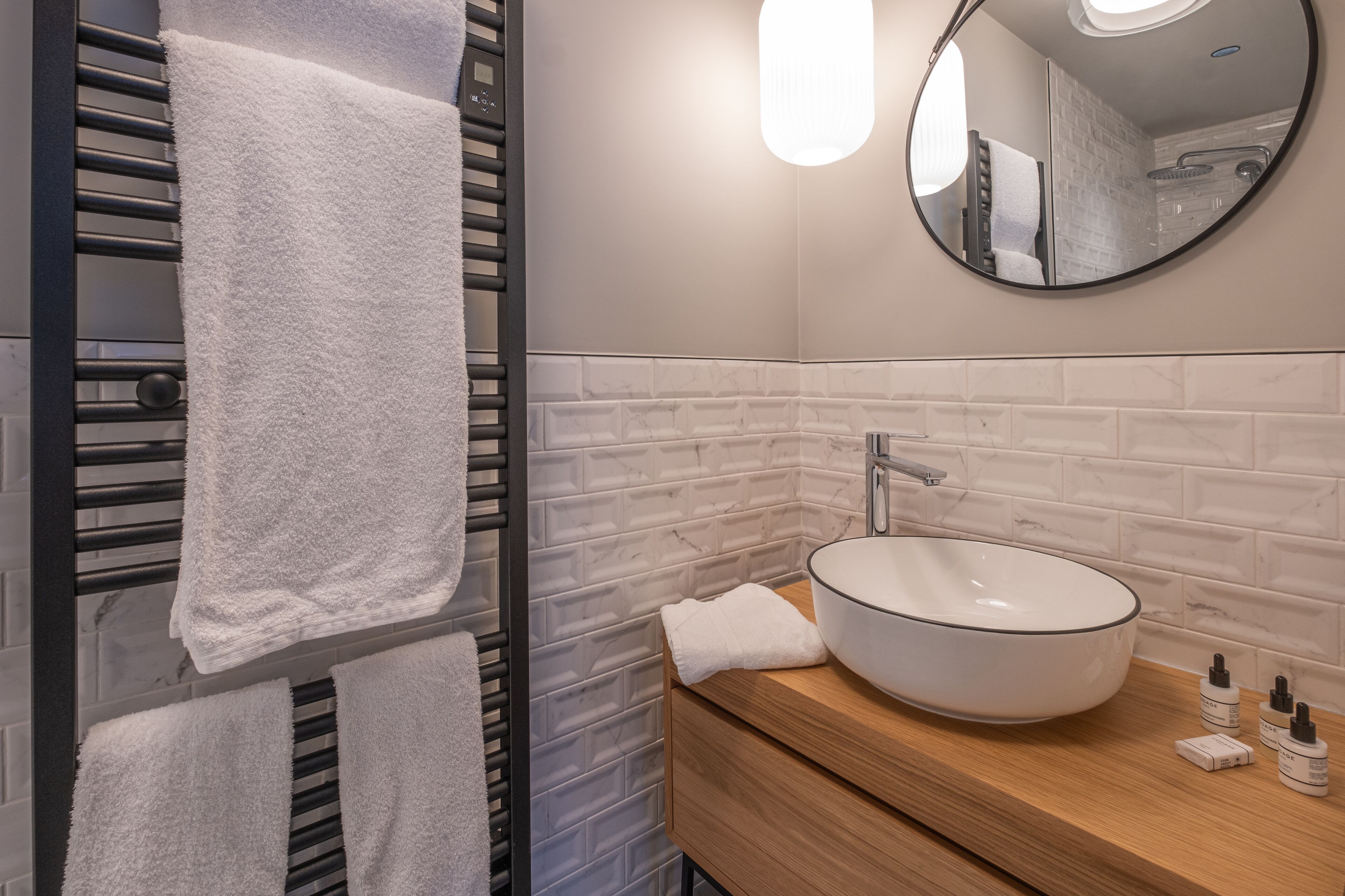 A modern bathroom with a towel rack holding two towels, a round mirror, a white sink with a wooden countertop, and a white tiled backsplash.