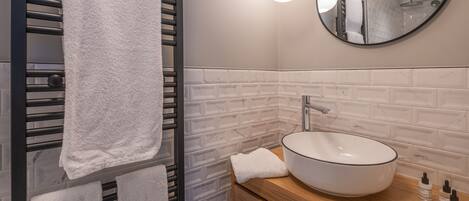 A modern bathroom with a towel rack holding two towels, a round mirror, a white sink with a wooden countertop, and a white tiled backsplash.