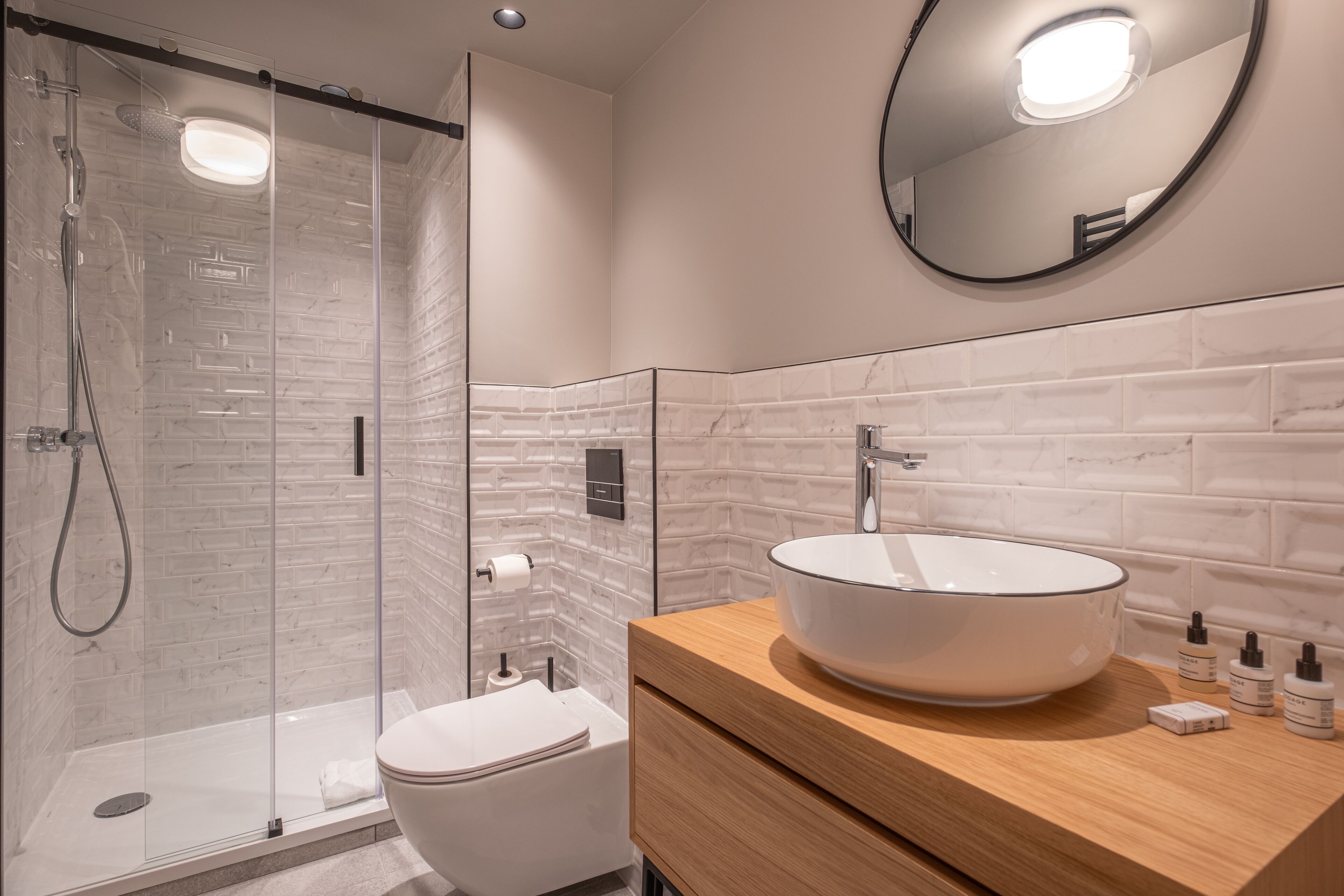 A modern bathroom with a glass shower enclosure, a white toilet, a wooden vanity with a large white sink, and a round mirror on the wall.