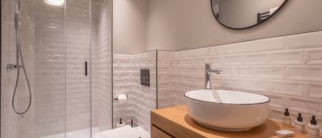 A modern bathroom with a glass shower enclosure, a white toilet, a wooden vanity with a large white sink, and a round mirror on the wall.