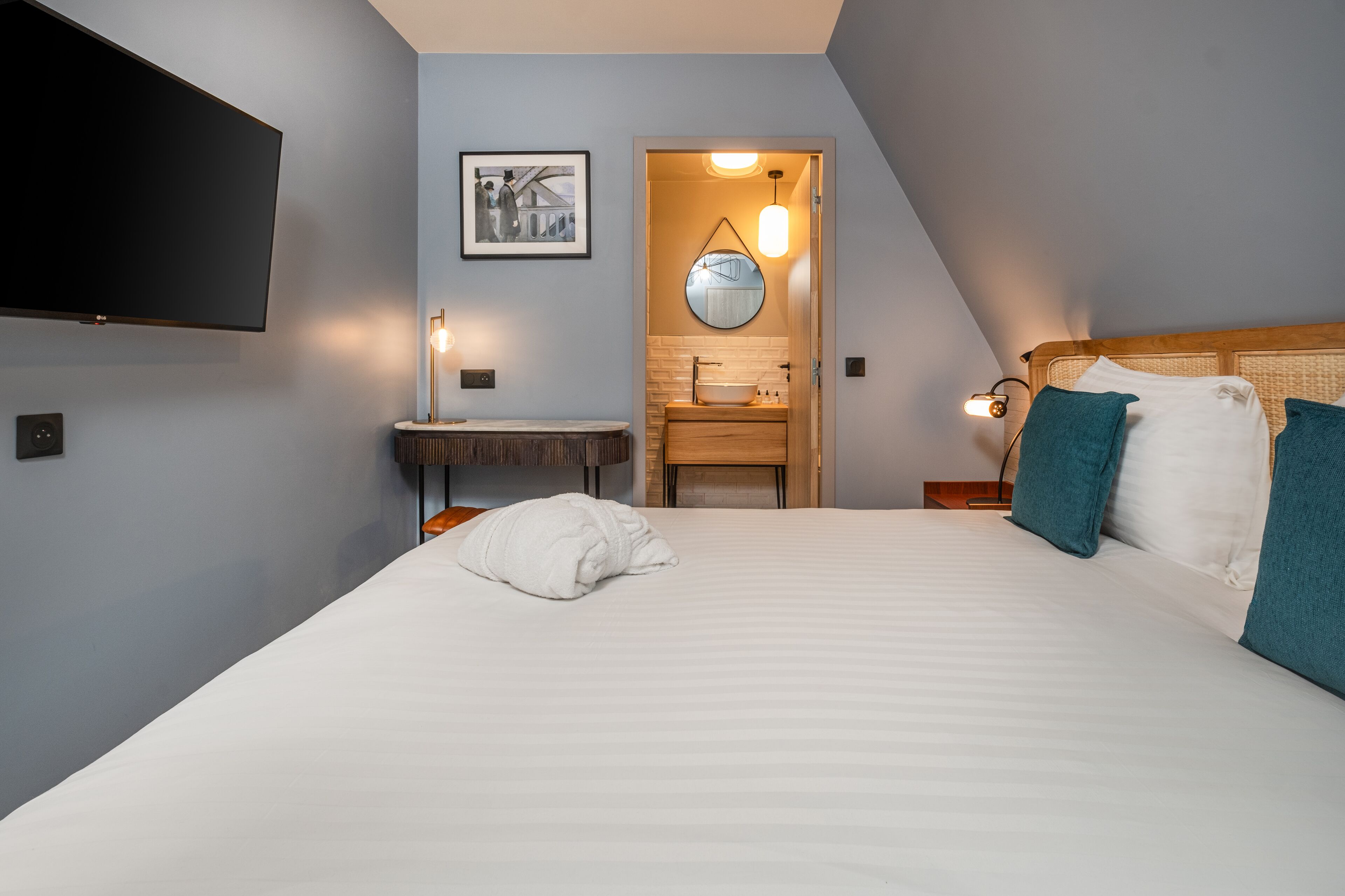 A modern hotel room with a large bed, a flat-screen TV, a small bathroom with a sink and mirror, and a framed picture on the wall.