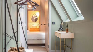 A narrow hallway with a sloped ceiling, a wooden bench with baskets, a hanging coat rack, and a desk with a lamp and a mirror. - Appart'hôtel East Paris Suite (Paris)