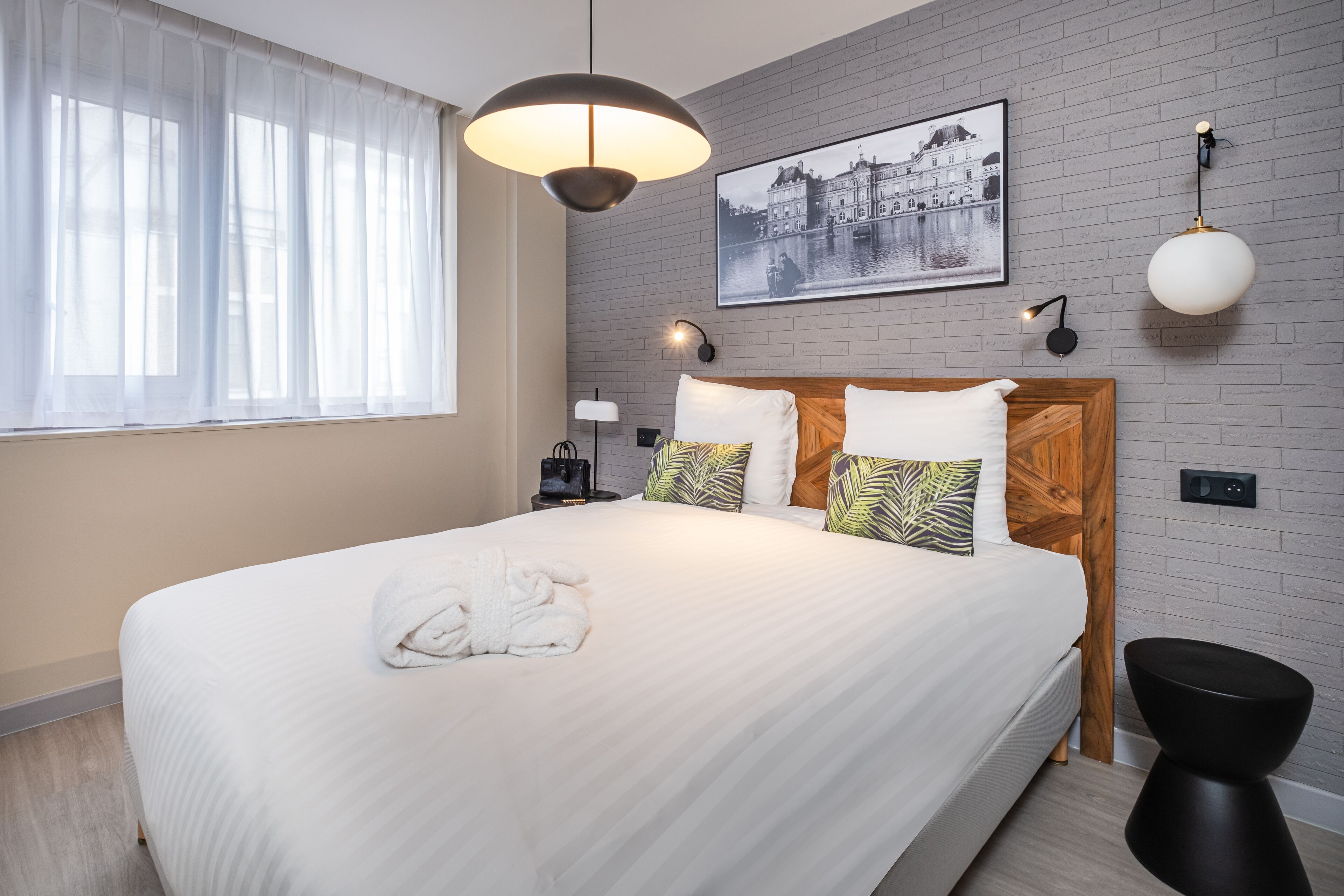 Een net opgemaakt bed met een witte deken en een opgevouwen handdoek, een houten hoofdbord, een zwart nachtkastje met een lamp, een aan de muur hangend schilderij en een ronde wandlamp.