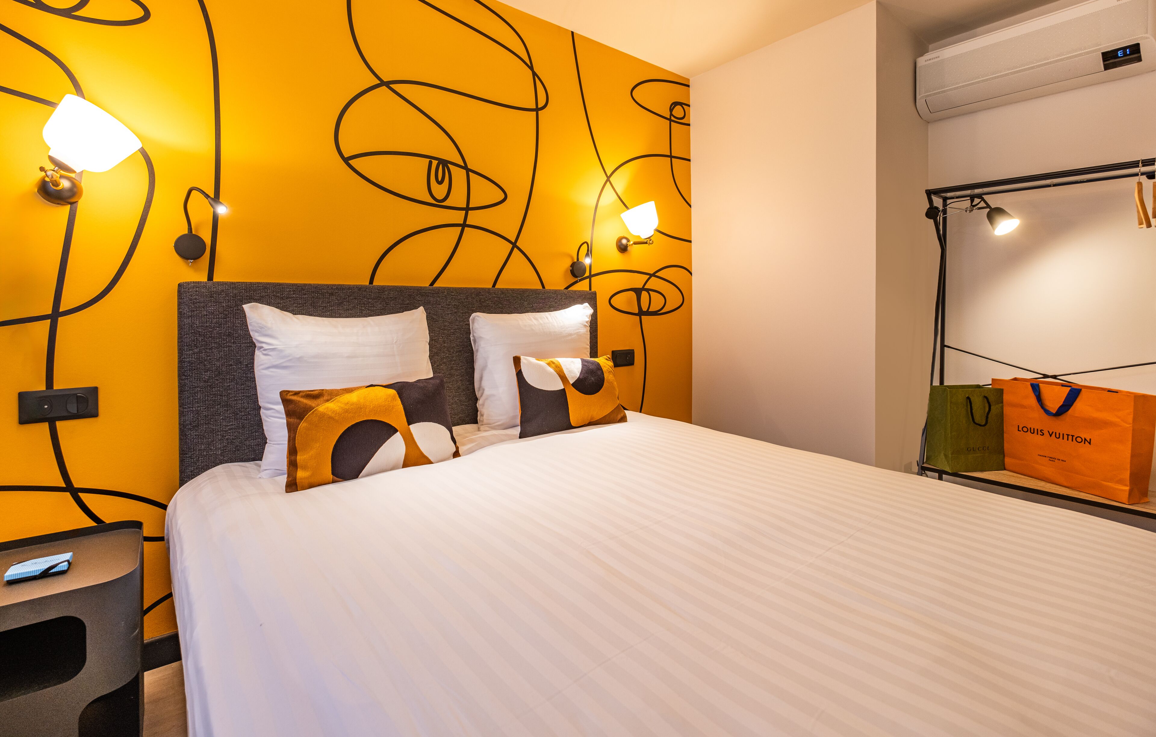 A hotel room with a bed, a nightstand, a wall-mounted lamp, and a luggage rack with bags.