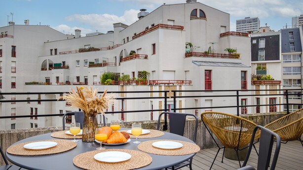 A rooftop terrace with a table set for breakfast, including croissants and orange juice, overlooking a white building with balconies. - Appart'hôtel East Paris Suite (Paris)