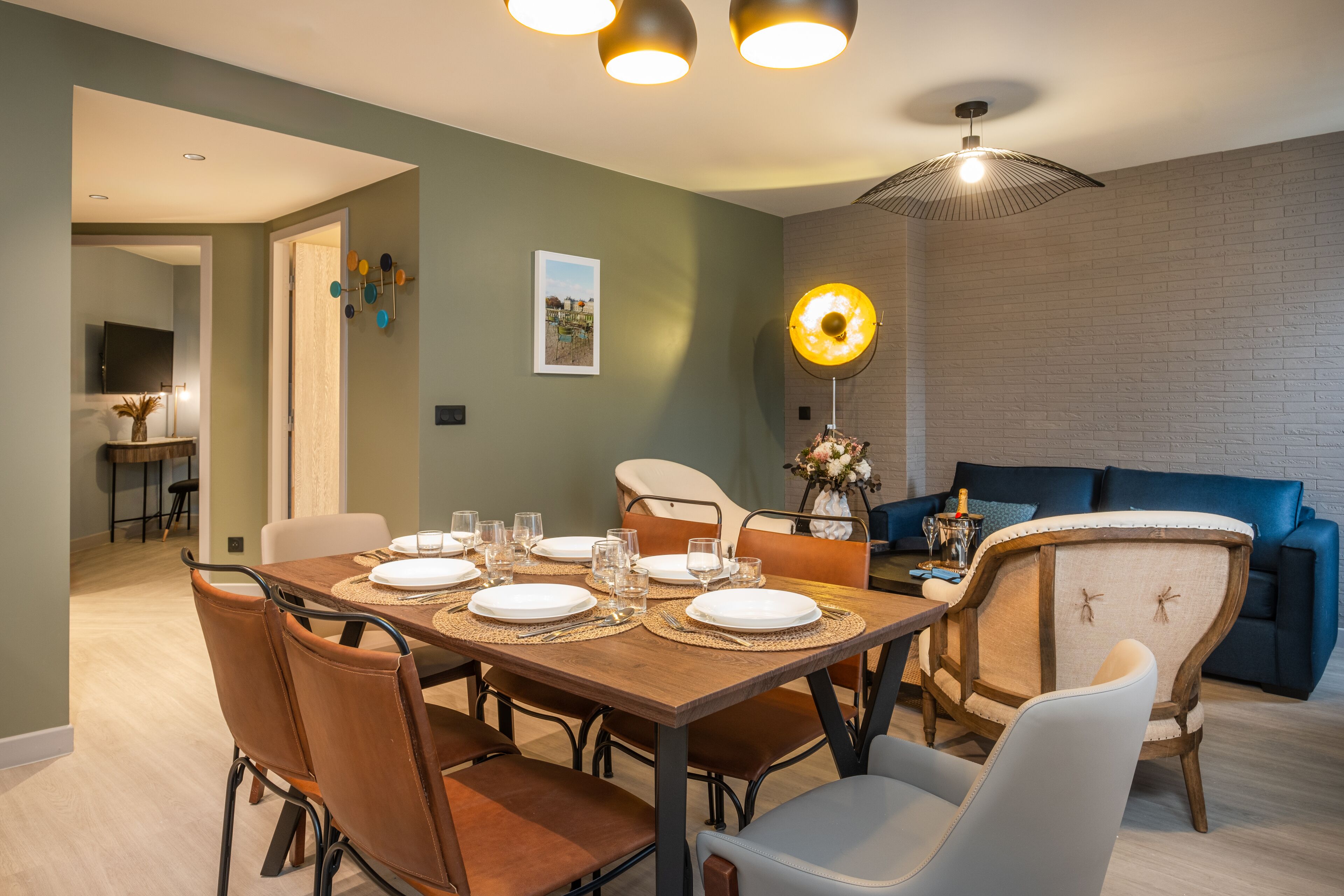 Un comedor con una mesa de madera dispuesta para seis personas, rodeada de sillas de cuero. Una lámpara colgante moderna ilumina la mesa, y se observa un sofá azul al fondo.