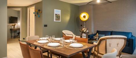 Un comedor con una mesa de madera dispuesta para seis personas, rodeada de sillas de cuero. Una lámpara colgante moderna ilumina la mesa, y se observa un sofá azul al fondo.