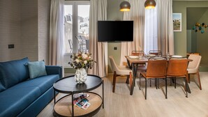 A modern living room with a blue sofa, a round coffee table with magazines, a dining area with a table and chairs, and a flat-screen TV mounted on the wall. - Appart'hôtel East Paris Suite (Paris)