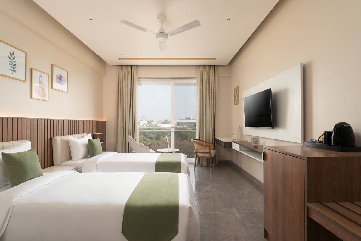 A hotel room with two beds, a TV, a desk, and a view of the city.