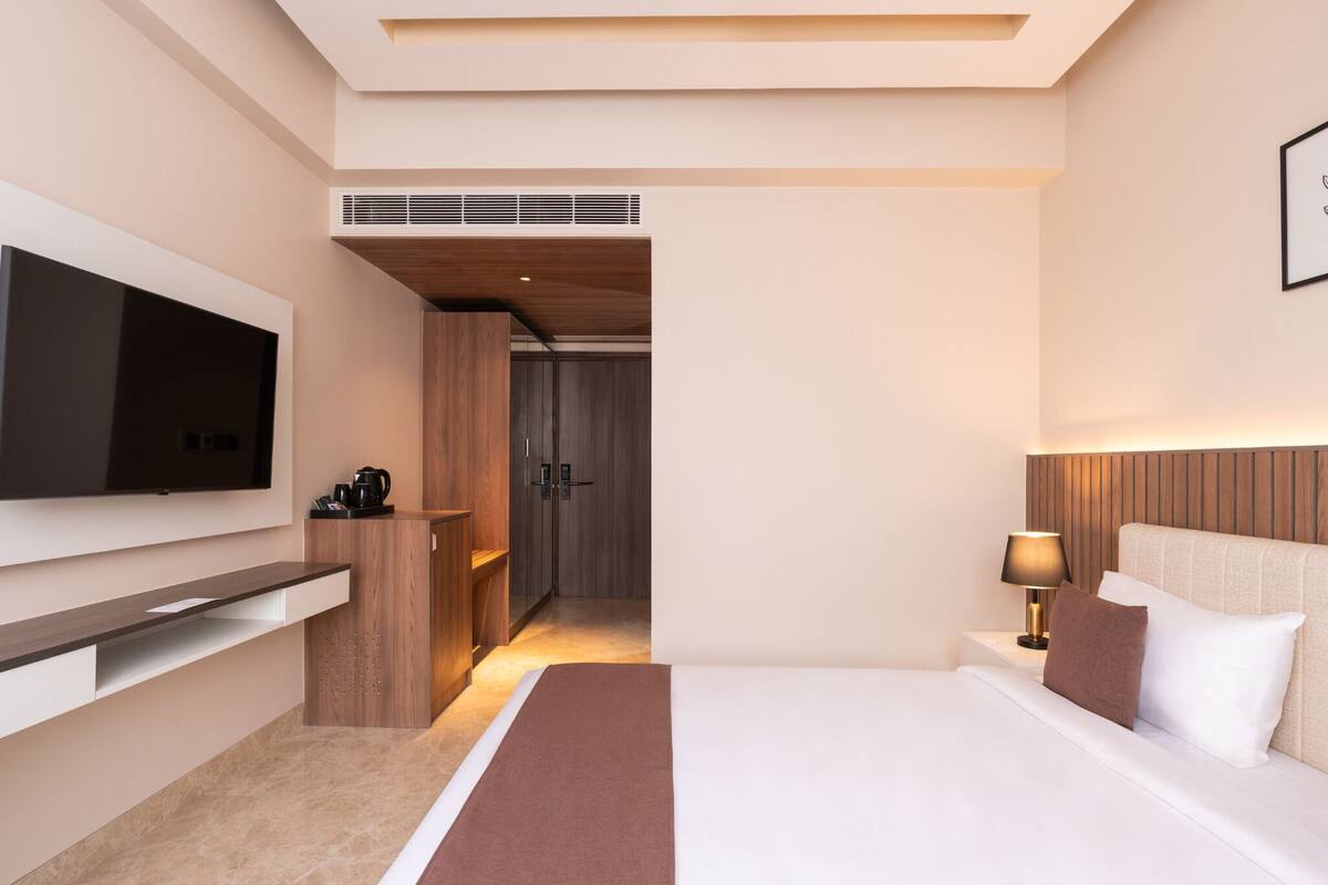 A modern hotel room with a large bed, a flat-screen TV mounted on the wall, a bedside table with a lamp, and a wooden wardrobe.