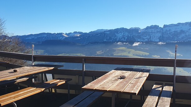 A terrace with wooden tables and benches overlooking a mountainous landscape. - Gertrud Schlössli (St. Gallen)