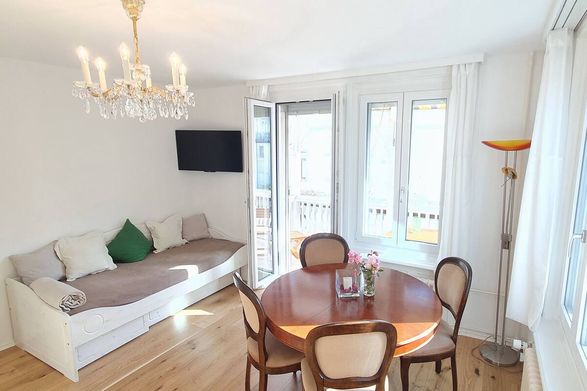 A modern living room with a wooden dining table, a sofa with cushions, a chandelier, and a TV mounted on the wall.