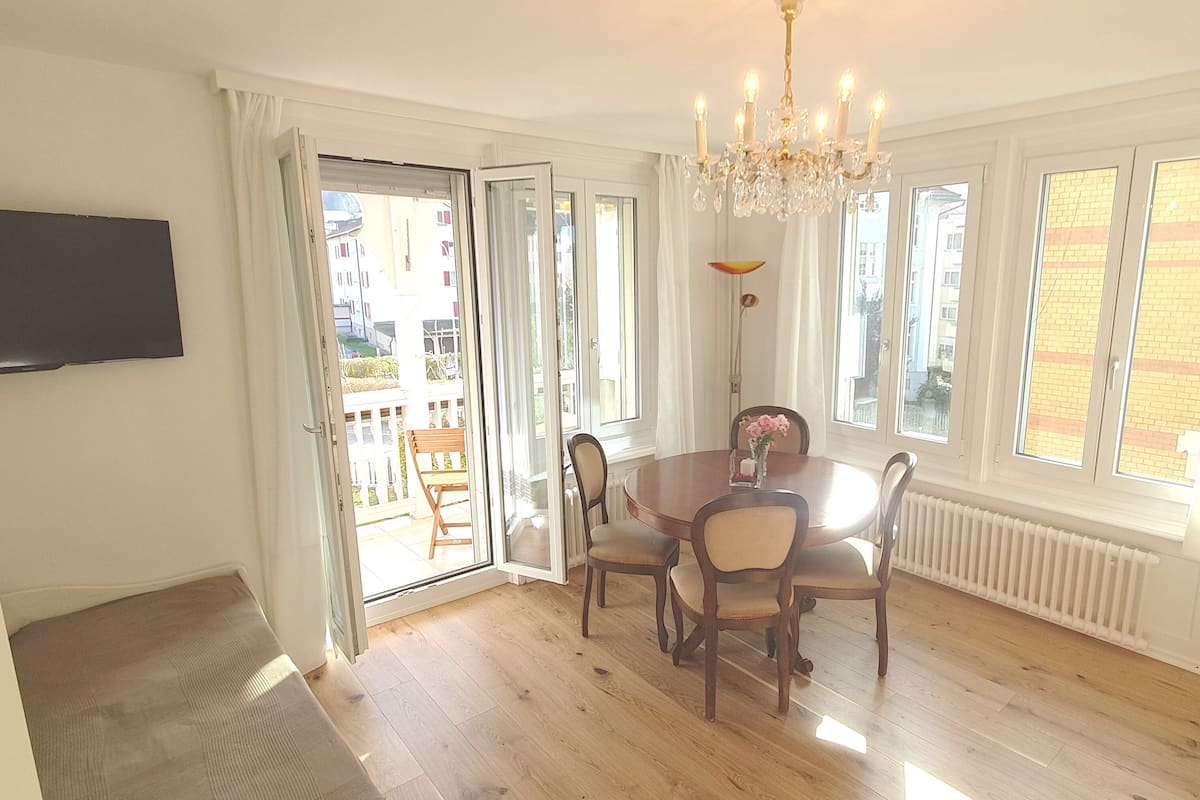 A dining area with a round table, chairs, and a chandelier. There is a television mounted on the wall and a window with a view of buildings.