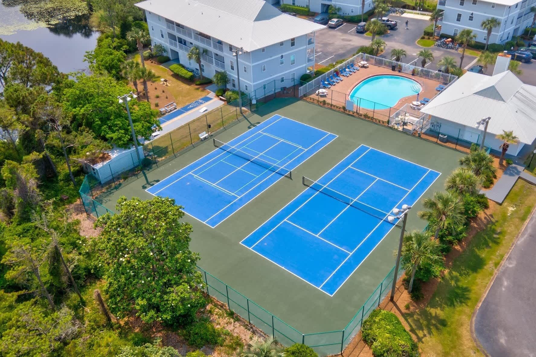 A tennis court surrounded by greenery and a pool, with a building in the background.